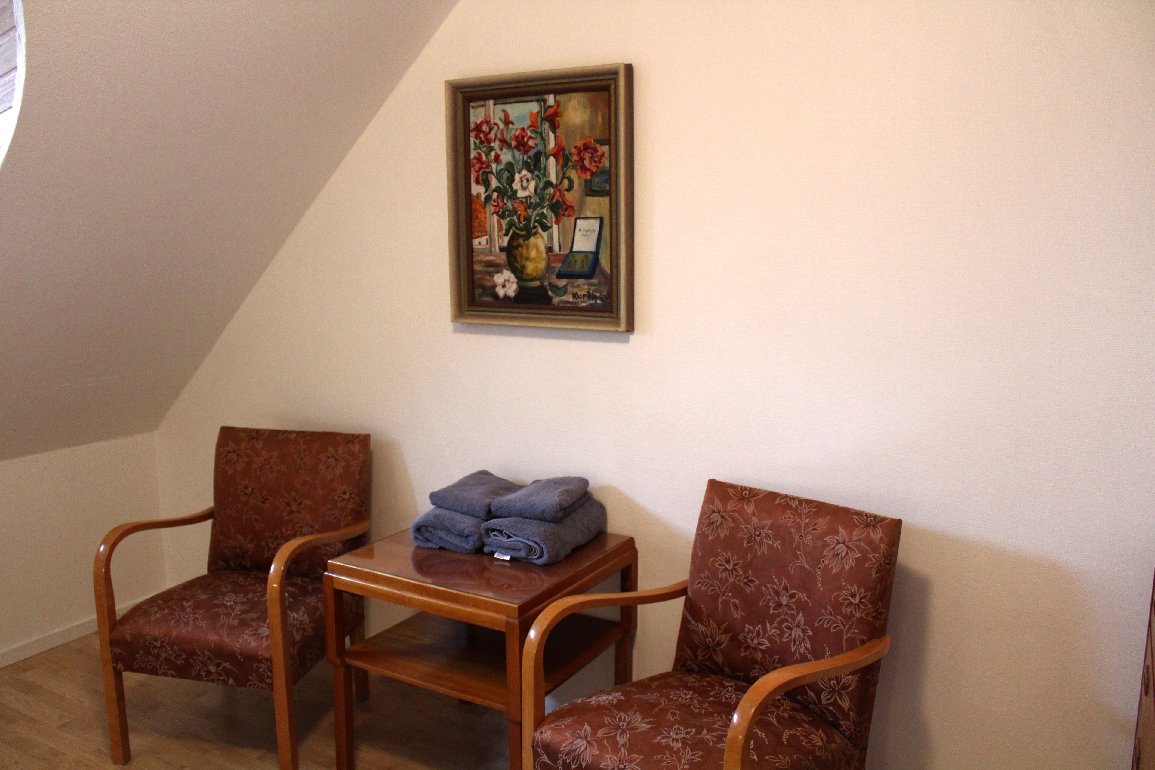 A room with two wooden chairs, a small table with folded towels, and a framed painting on the wall.
