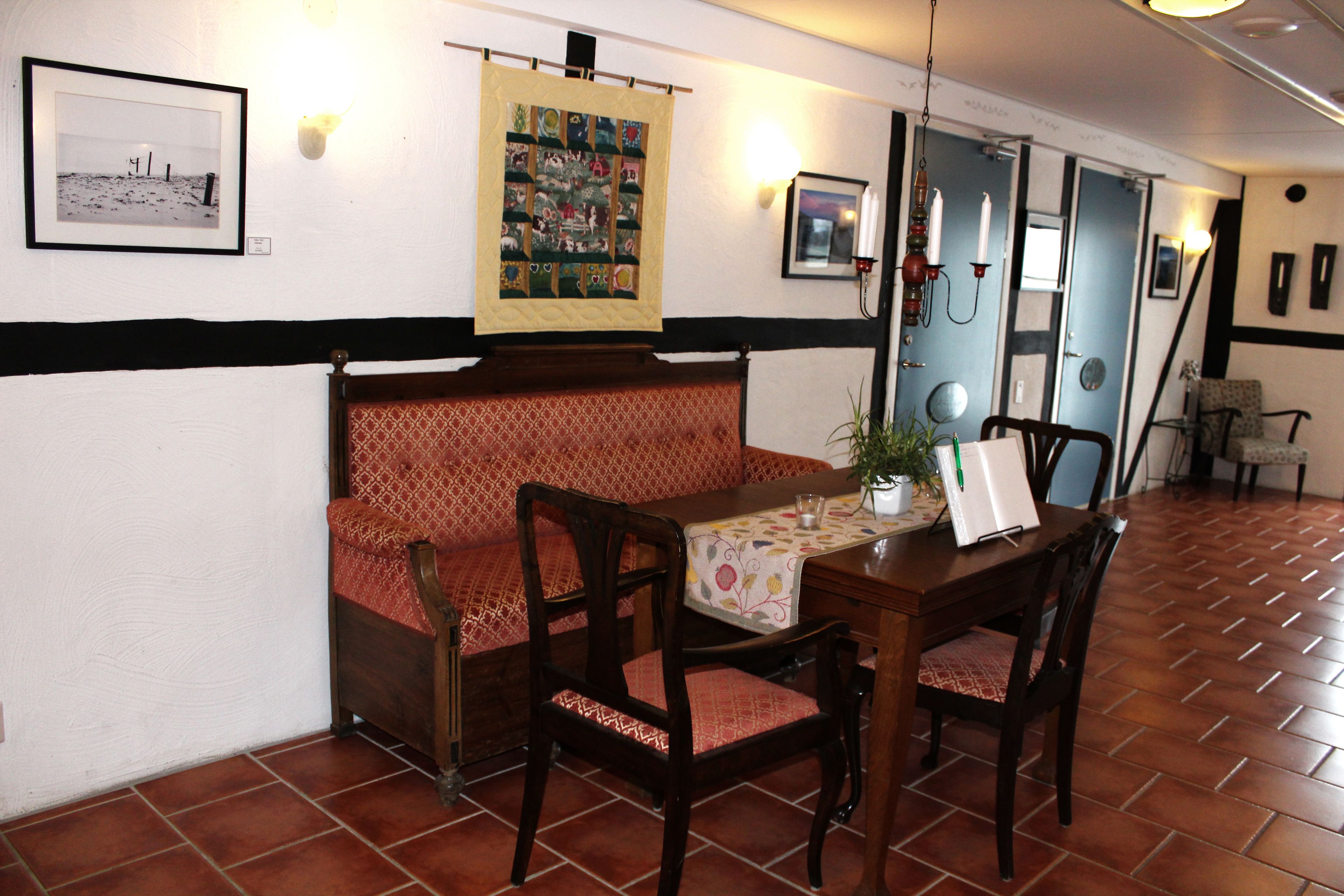 A dining area with a wooden table, chairs, and a bench. There is a quilt hanging on the wall and a framed picture above the bench.