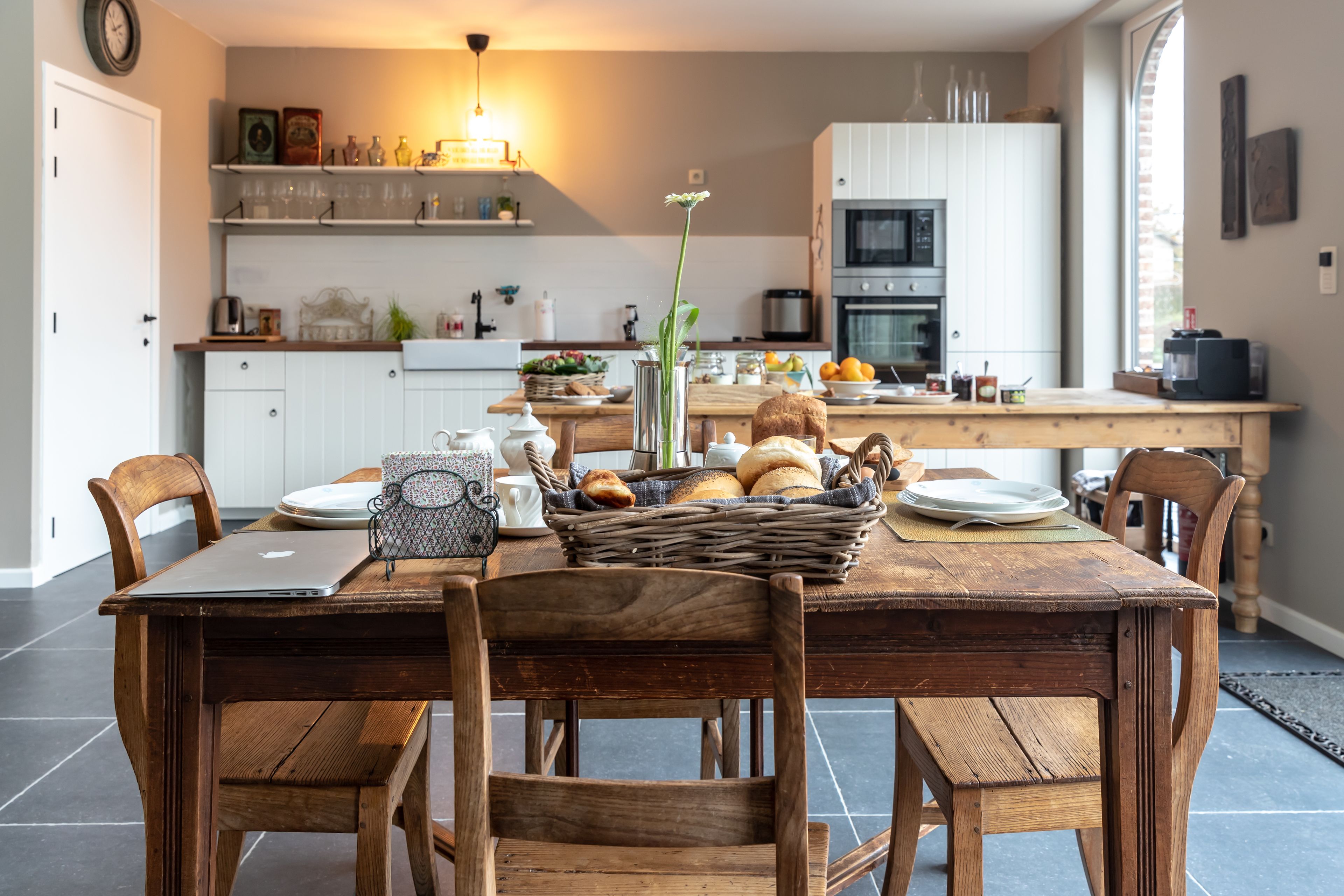 Un comedor con mesa de madera, sillas y una computadora portátil. En el fondo, una cocina con armarios, un microondas y un fregadero.