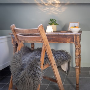 Una silla de madera con asiento y respaldo de piel, una mesa de madera con un cuenco de fruta, una planta pequeña y un cuadro enmarco.
