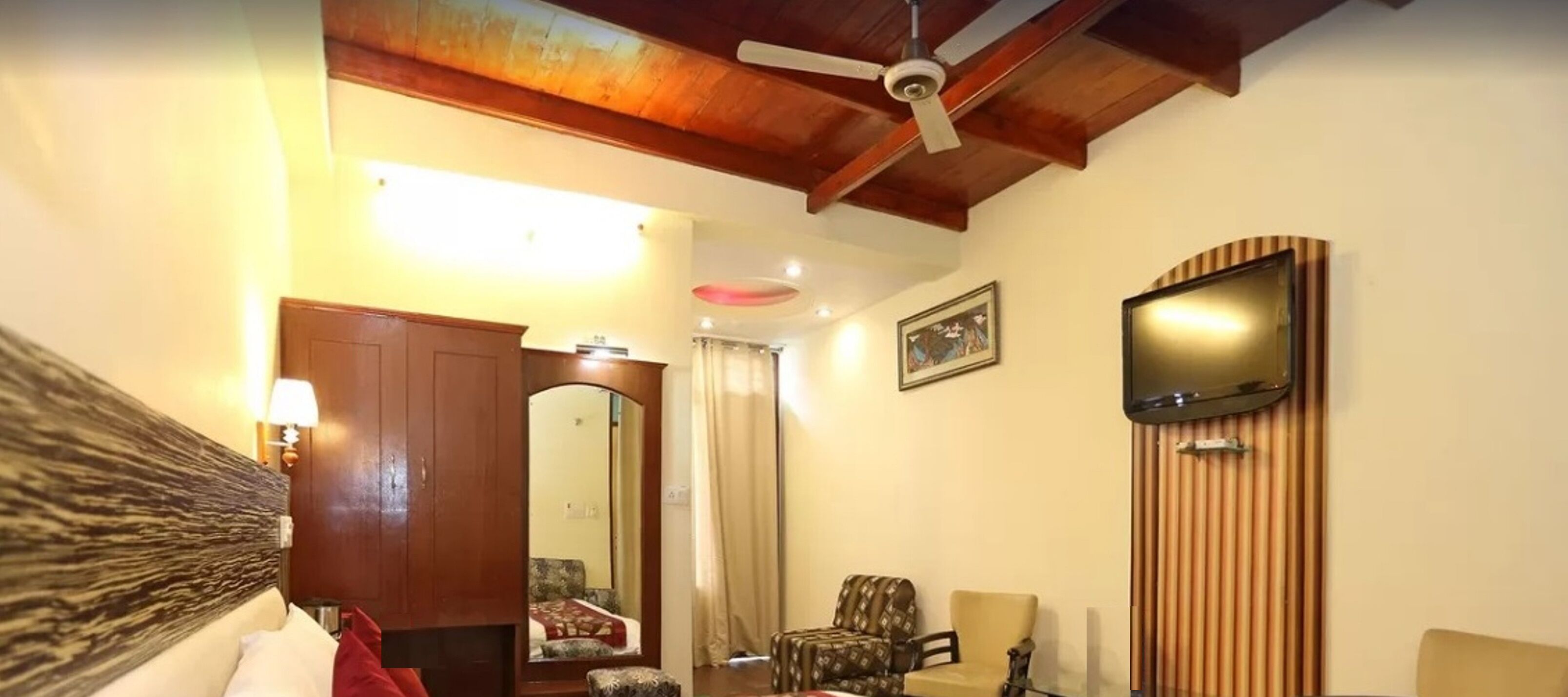 A room with a wooden ceiling, a television mounted on the wall, a bed with a headboard, a chair, and a mirror.