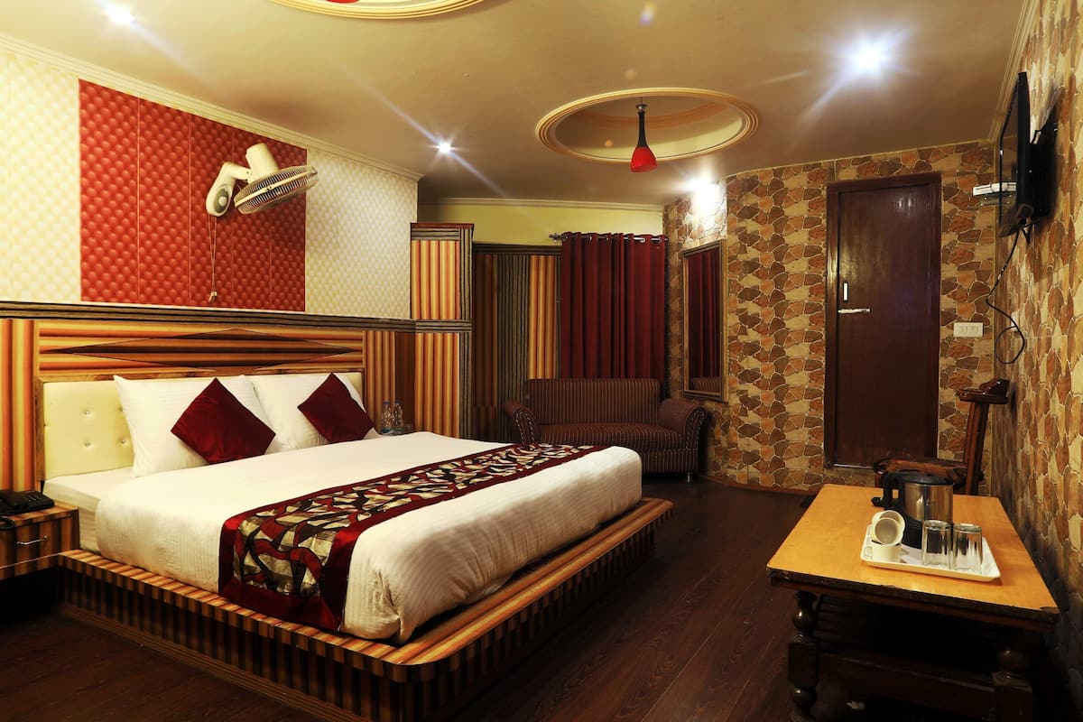 A hotel room with a bed, a wooden bench, a table with a coffee pot and glasses, and a TV mounted on the wall.