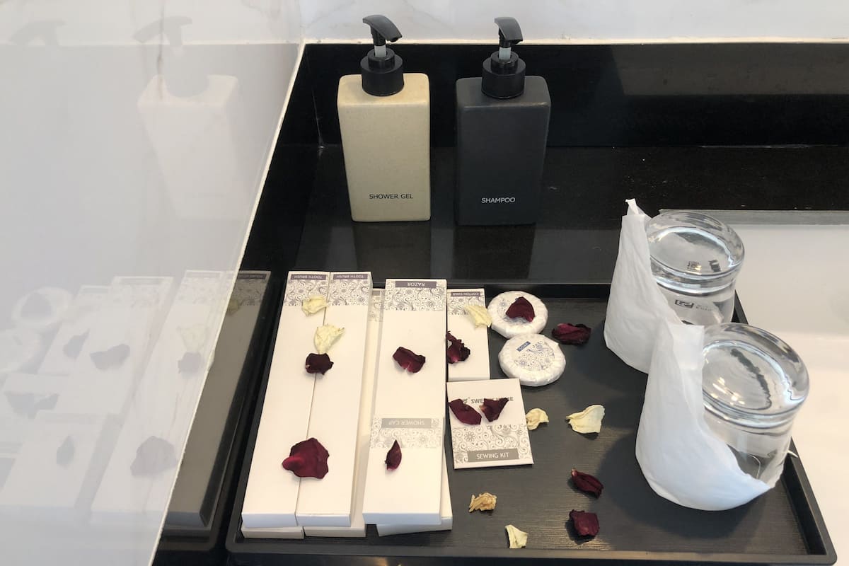 A bathroom counter with a black tray holding various toiletries, including a shower gel bottle, a shampoo bottle, and a sewing kit.