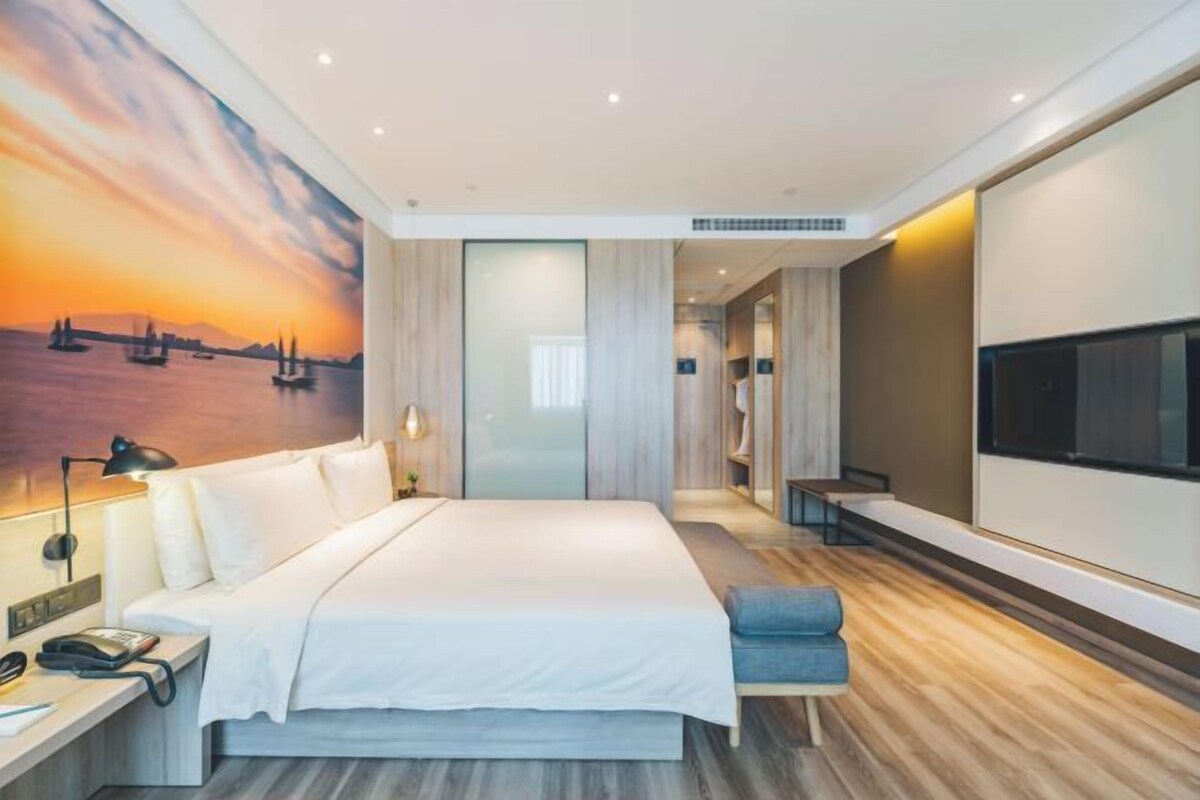 A modern hotel room with a large bed, a TV, and a painting of a sunset over water.