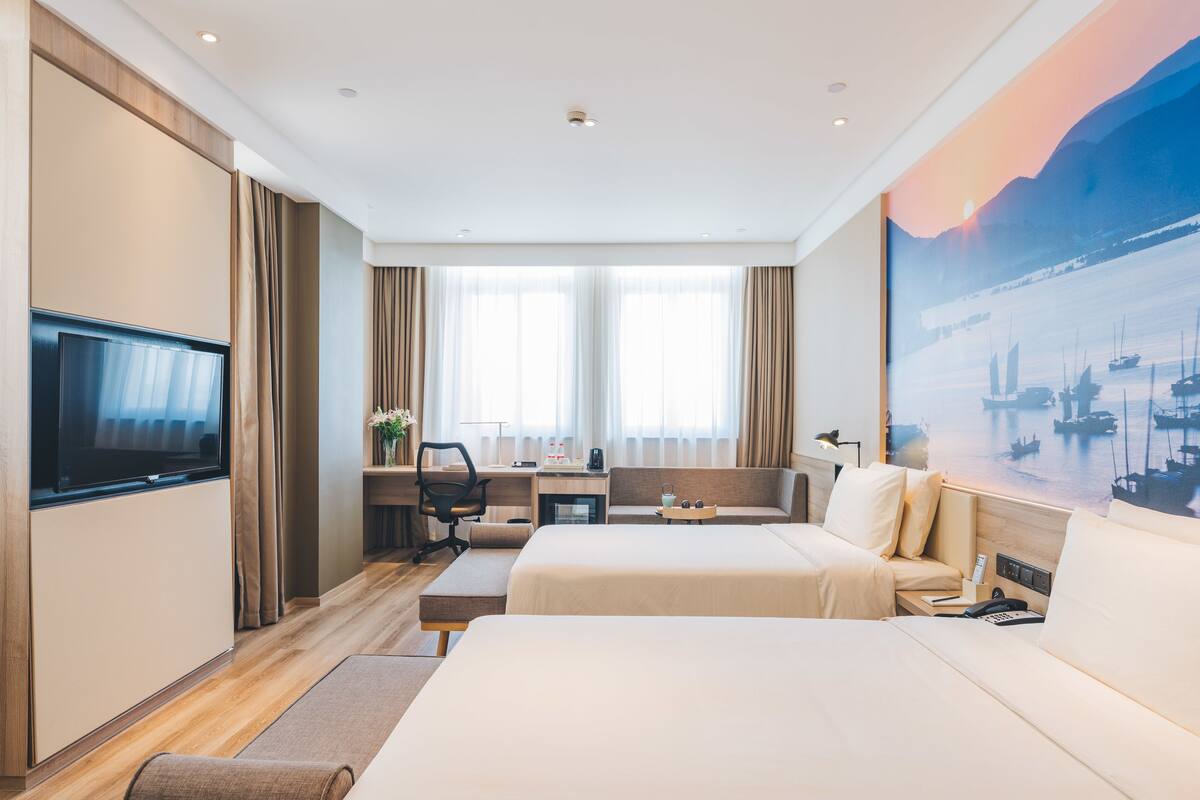 A hotel room with two beds, a desk with a chair, a TV, and a painting of a harbor scene.