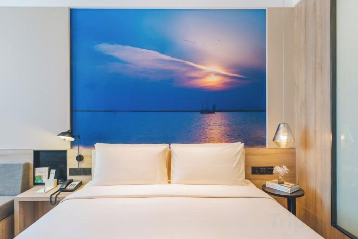 A hotel room with a bed, a nightstand, a lamp, and a wall-mounted painting of a sunset over water.