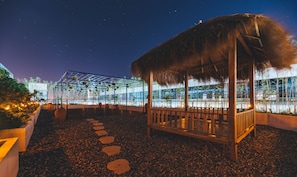 A night scene of a resort with a thatched-roof gazebo, a glass-enclosed structure, and a pathway of stepping stones.