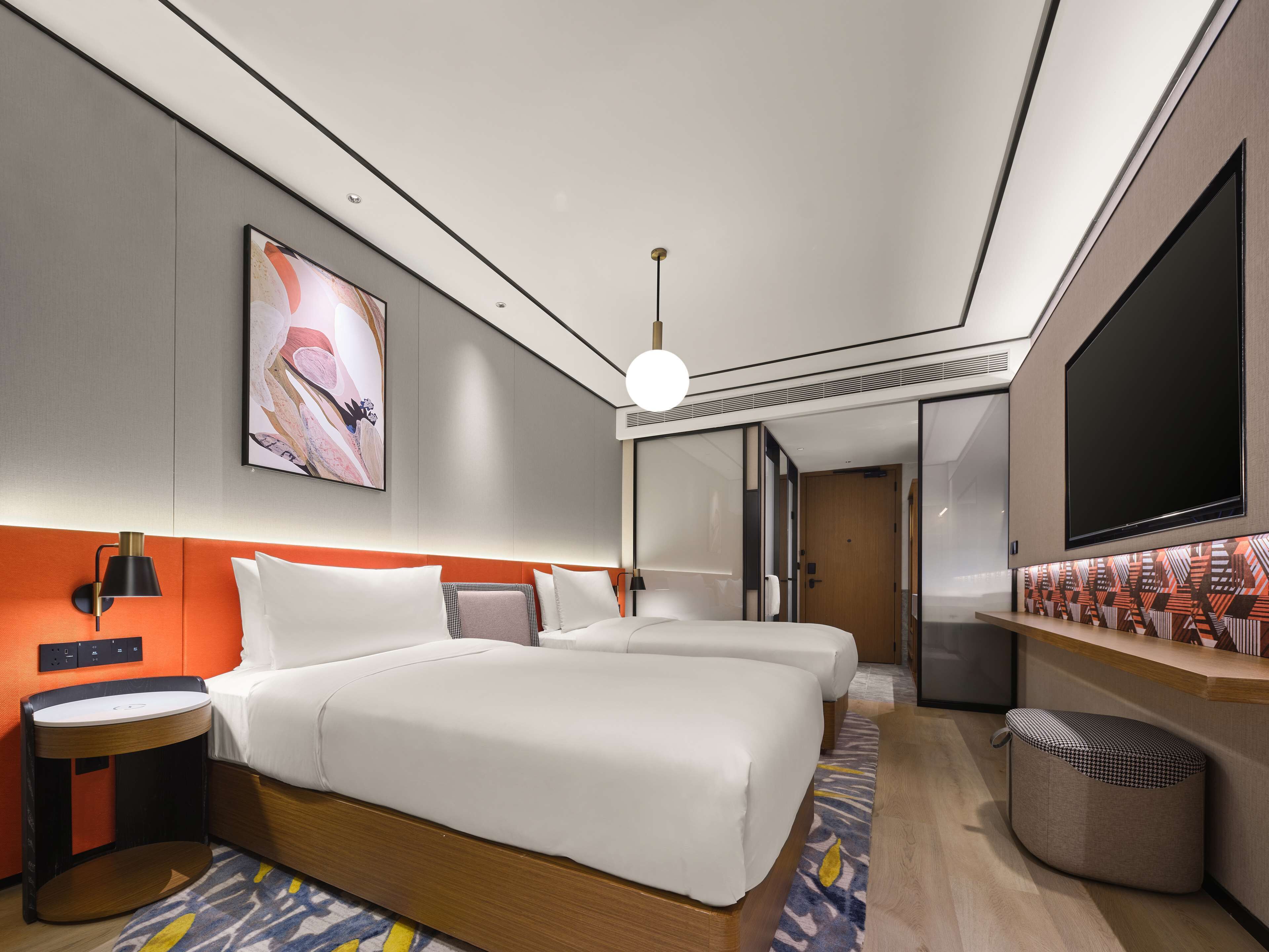 Habitación de hotel moderna con dos camas, un televisor, un escritorio y un baño visible a través de una puerta de cristal.