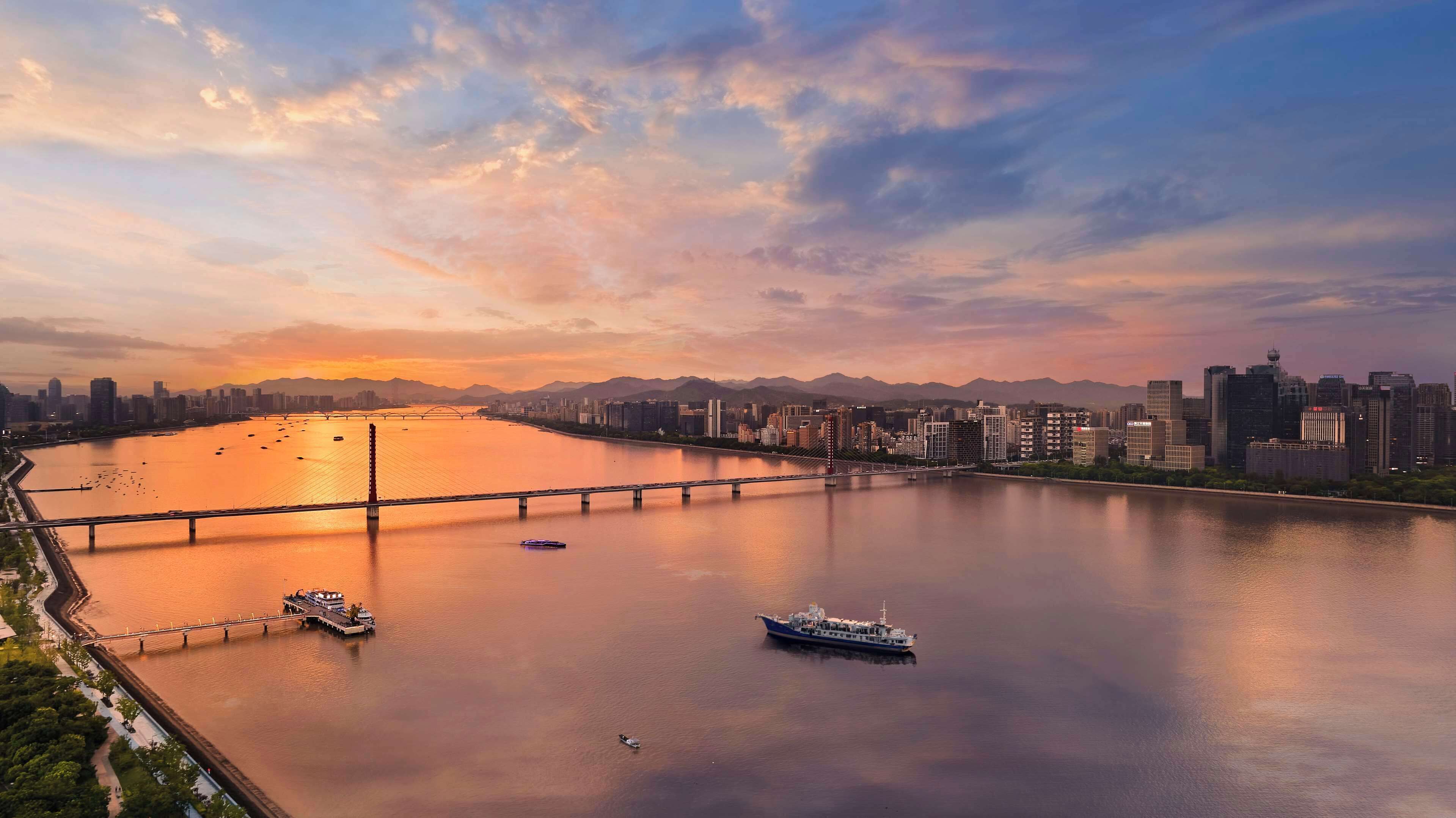 Un río con un puente y barcos, un horizonte urbano al fondo, y montañas a lo lejos.