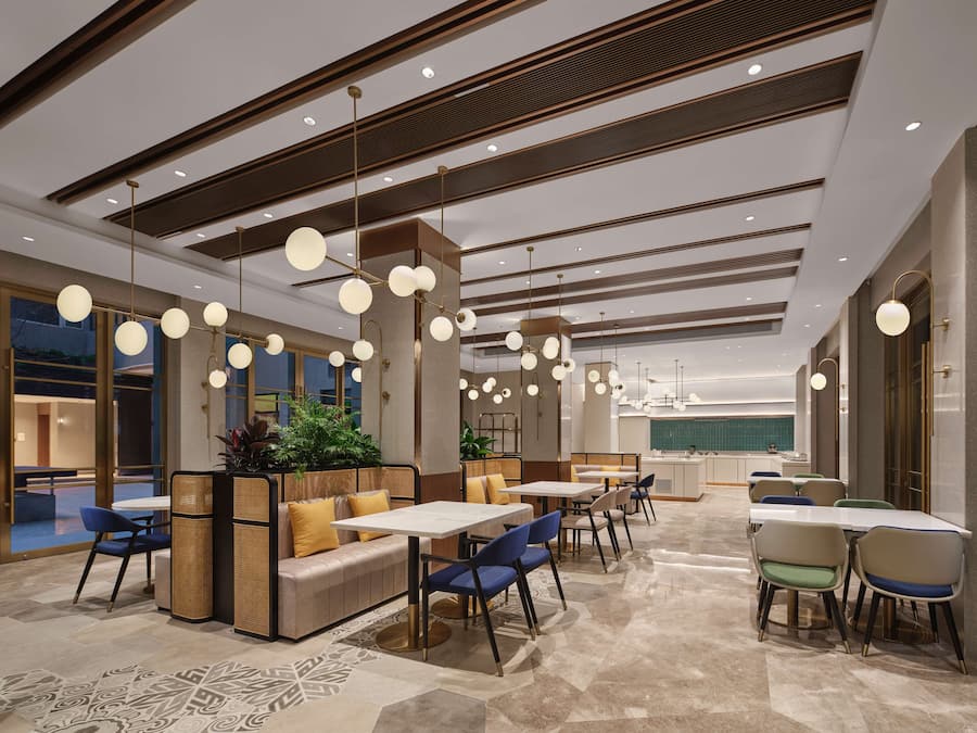 Um hall de hotel moderno com áreas de estar, mesas e uma cozinha ao fundo.