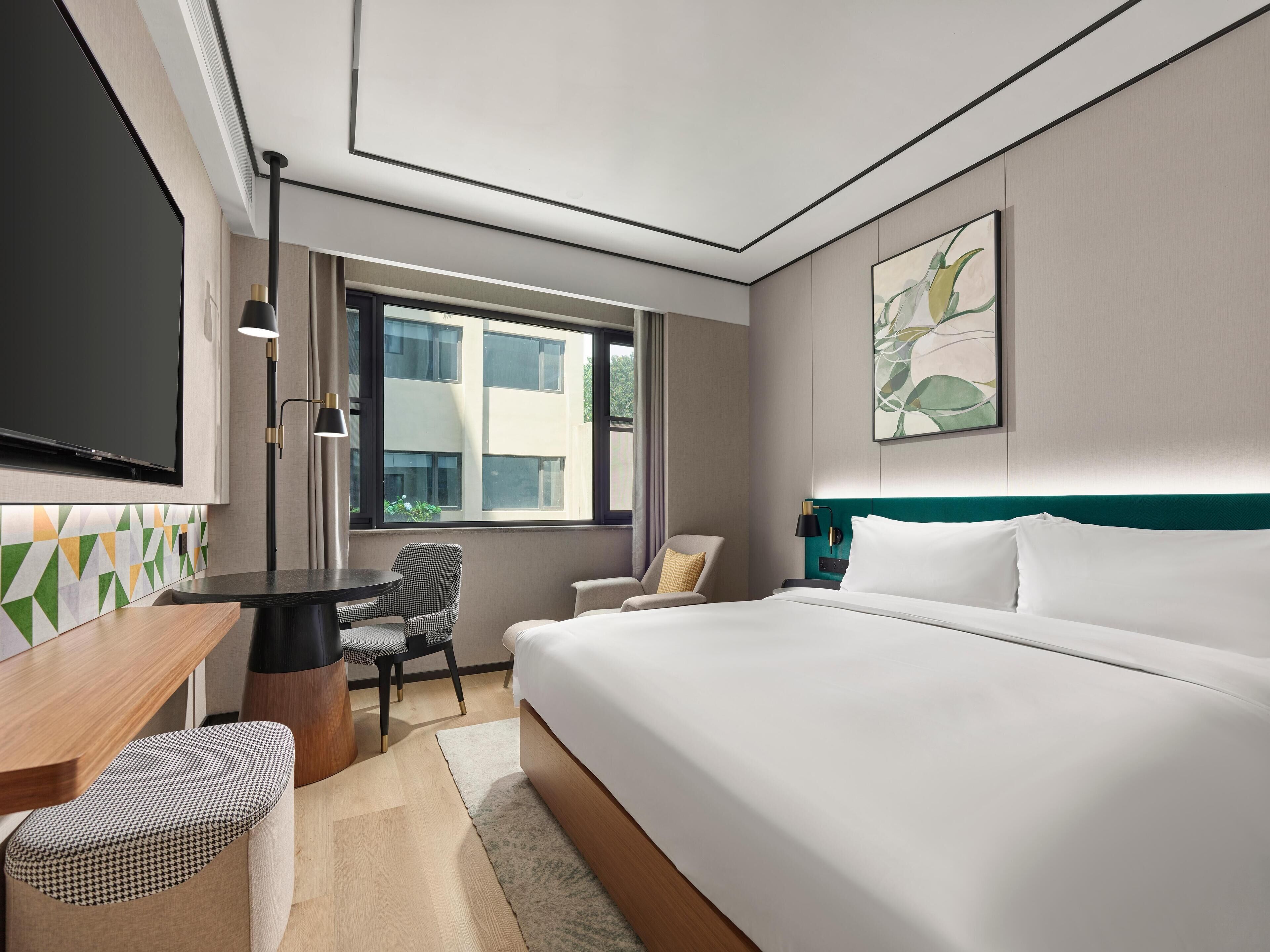 A hotel room with a large bed, a desk with a chair, a TV, and a window with a view of buildings.