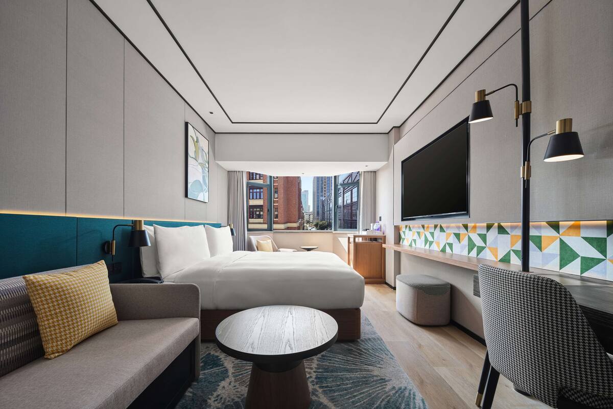 Una habitación de hotel moderna con cama, sofá y una pequeña mesa. Hay un televisor montado en la pared y una ventana con vistas a la ciudad.