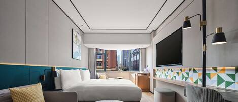 Una habitación de hotel moderna con cama, sofá y una pequeña mesa. Hay un televisor montado en la pared y una ventana con vistas a la ciudad.