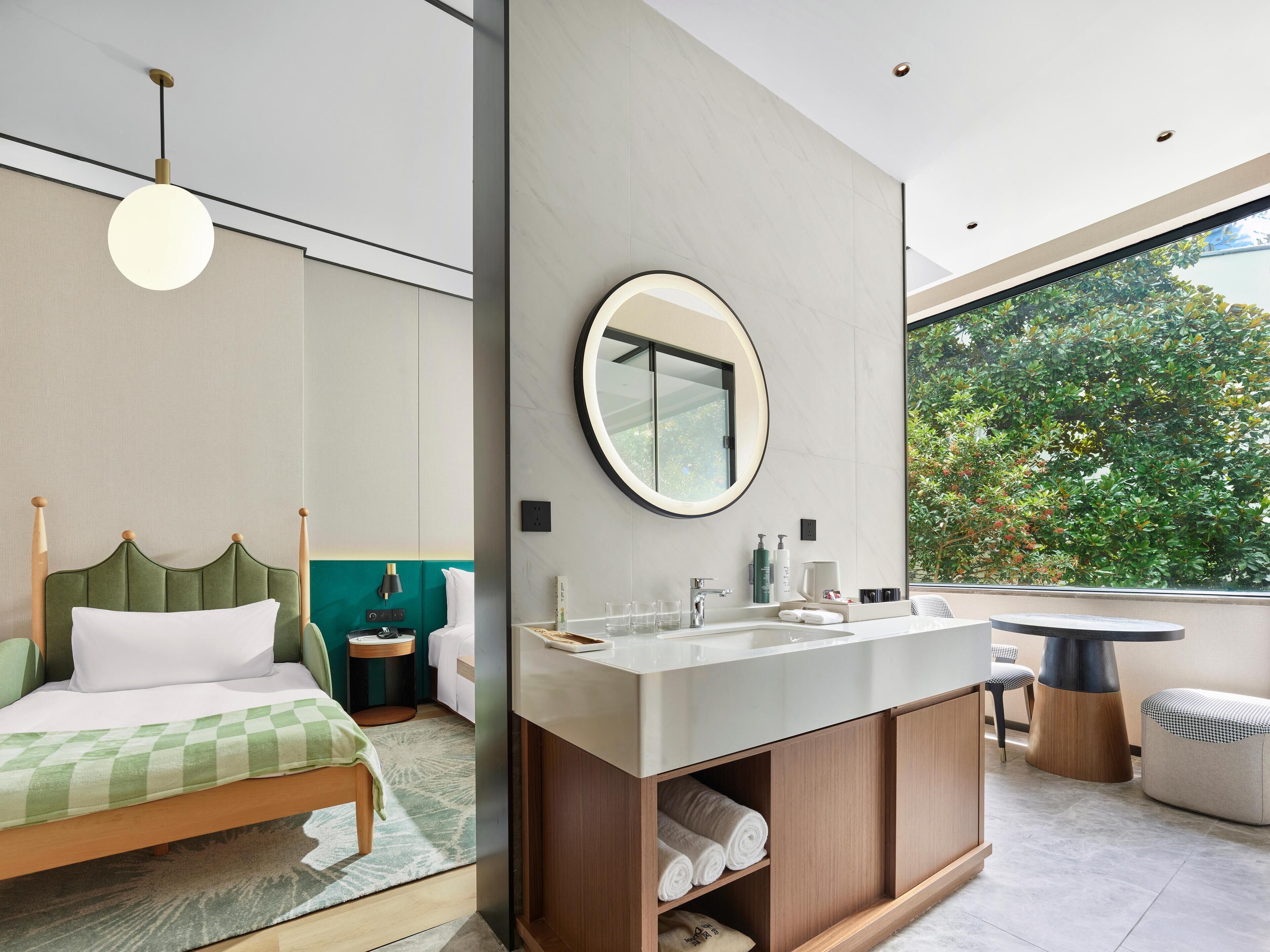 Quarto moderno com cama, mesa de cabeceira, espelho redondo, banheiro com pia e vista para o verde lá fora.
