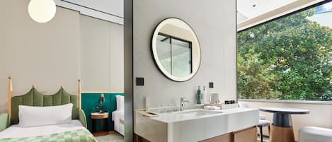 A modern bedroom with a bed, a bedside table, a round mirror, a bathroom with a sink, and a view of greenery outside.