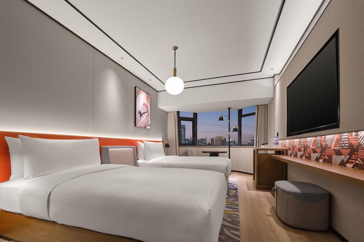 Habitación de hotel moderna con dos camas, un televisor grande y vista a la ciudad.