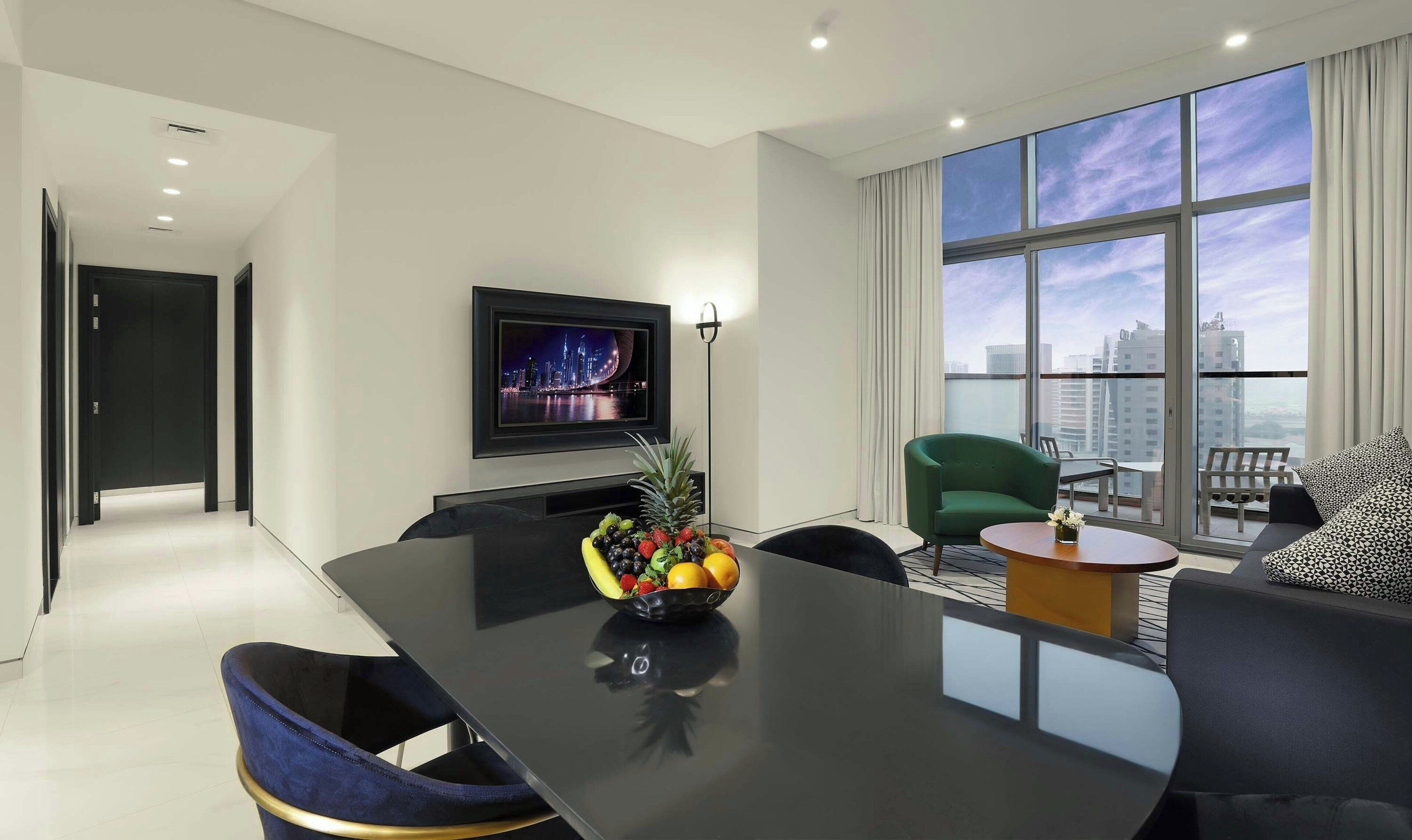 A modern hotel room with a large window, a flat-screen TV, a dining area, and a fruit bowl on the table.