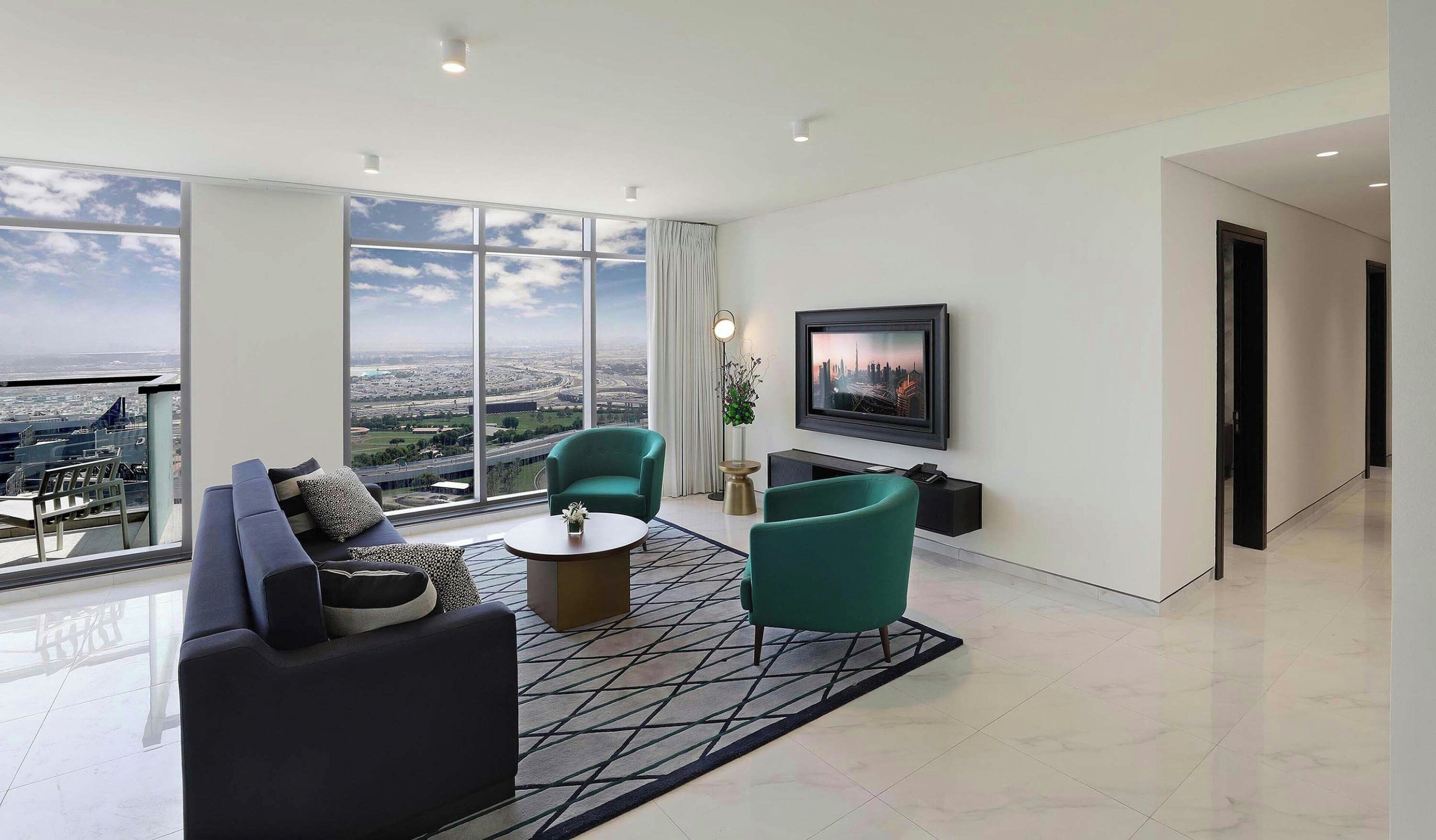 A modern living room with a large window, a sofa, two armchairs, a coffee table, and a flat-screen TV mounted on the wall.