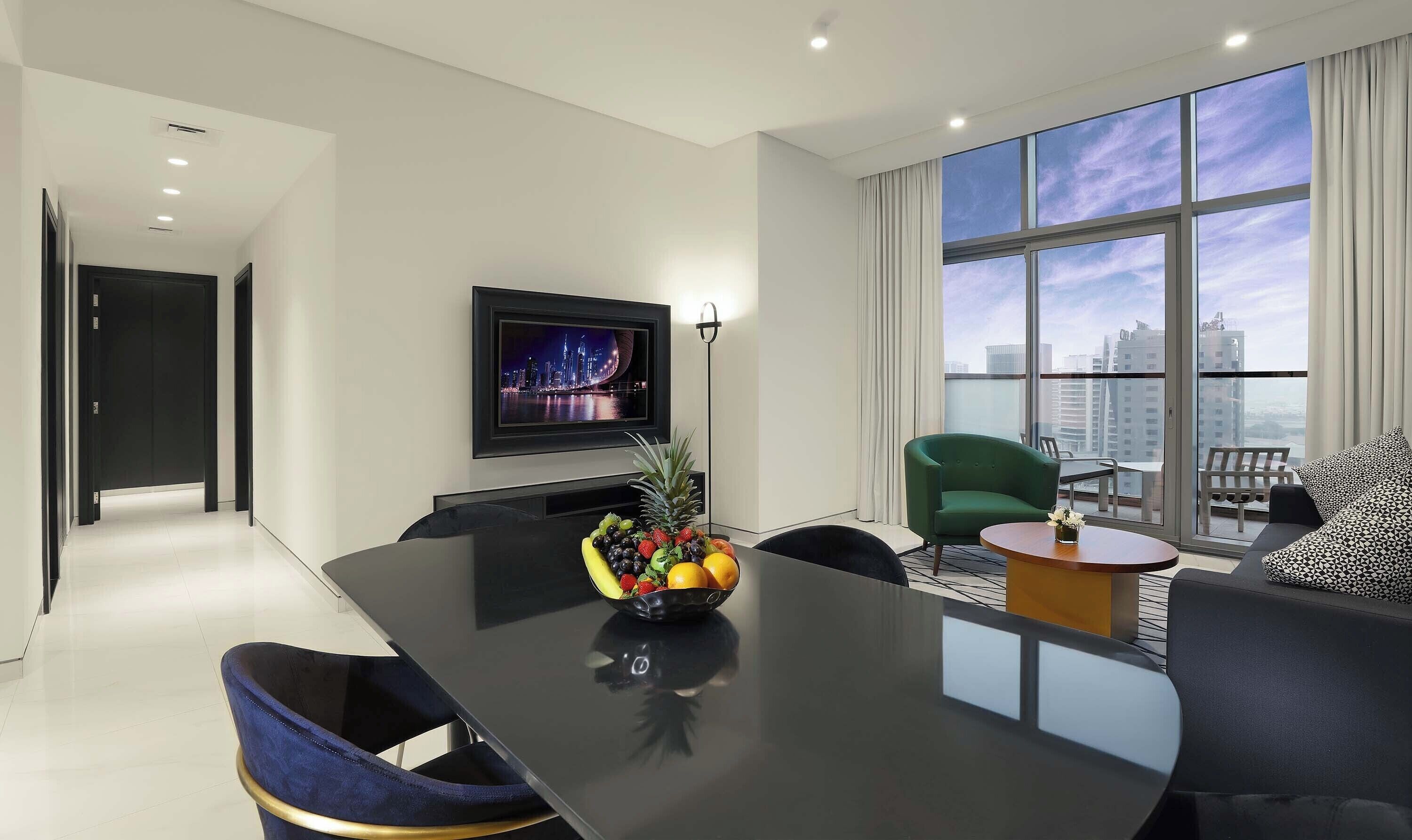 A modern hotel room with a large window, a flat-screen TV, a dining table with chairs, and a fruit bowl.