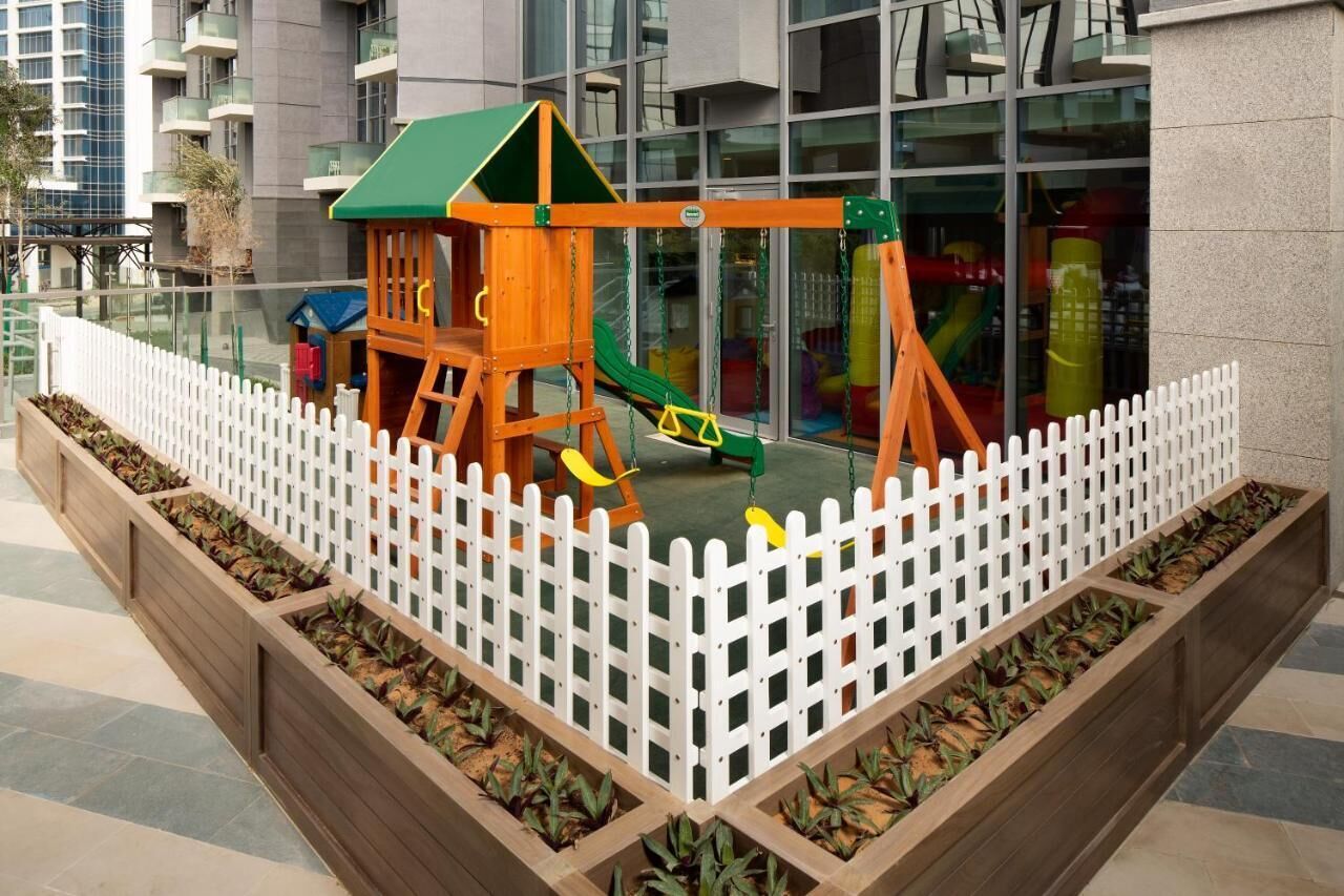 A children's play area with a swing set and slide, surrounded by a white picket fence and wooden planters.