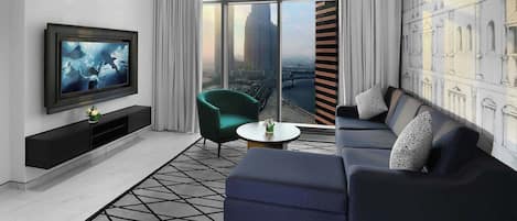 A modern living room with a large TV, a green armchair, a sofa with patterned cushions, and a city view through floor-to-ceiling windows.