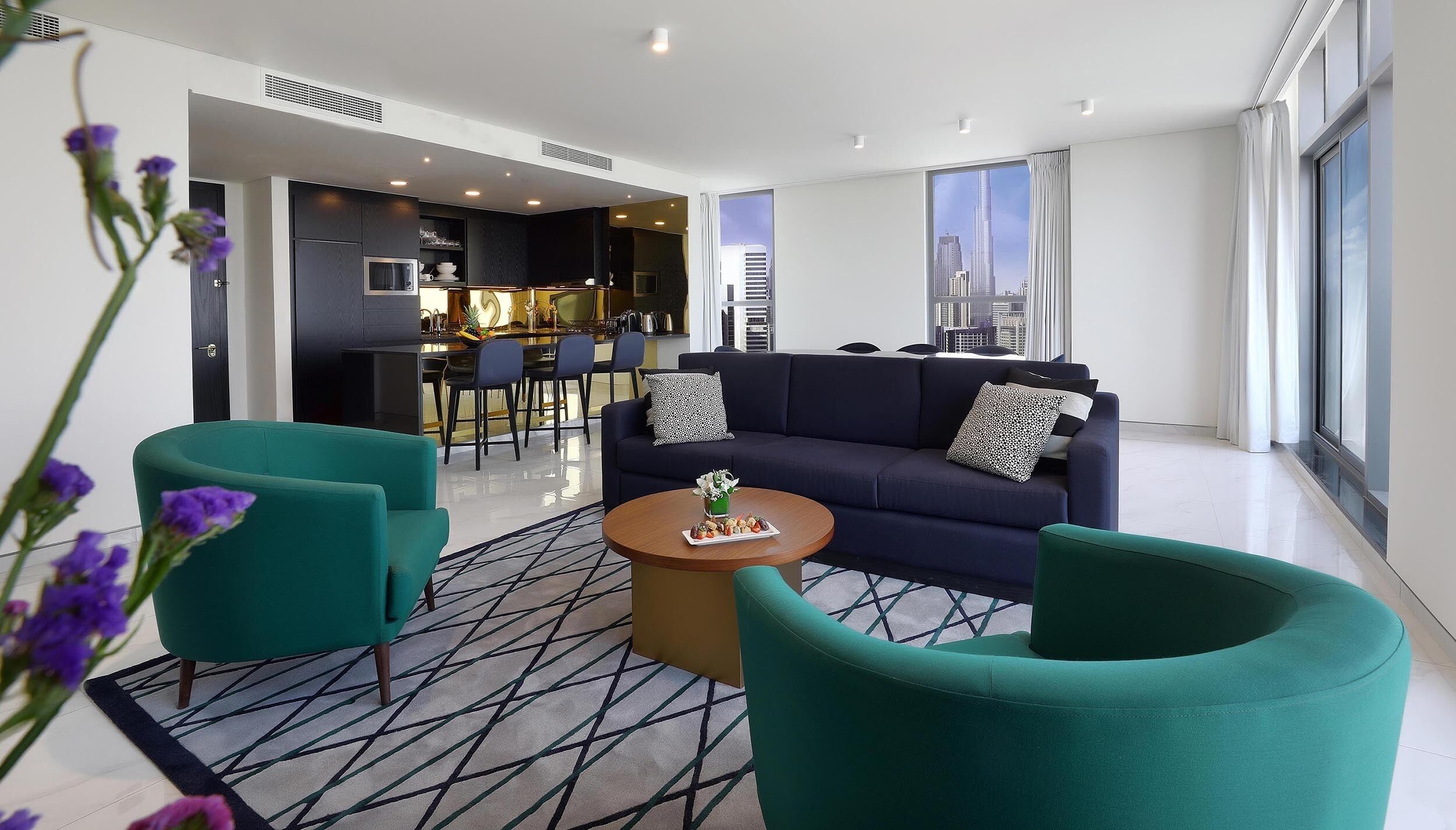 A modern living room with teal armchairs, a dark sofa, and a small round table with snacks. A kitchen is visible in the background with a city view through large windows.