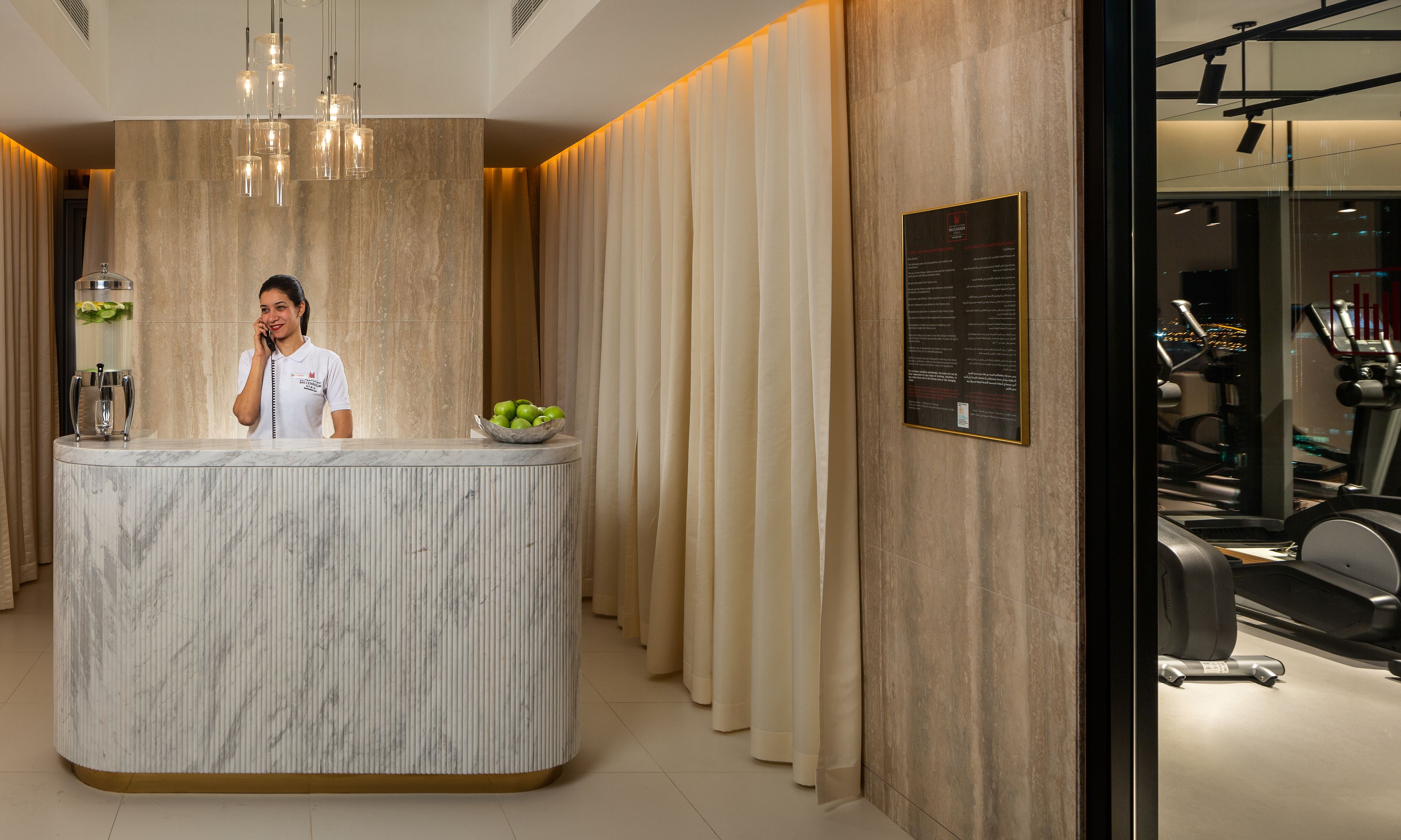 A reception desk with a marble finish, a water dispenser, and a bowl of green apples. There is a wall-mounted sign with text and a view of a gym with exercise equipment.