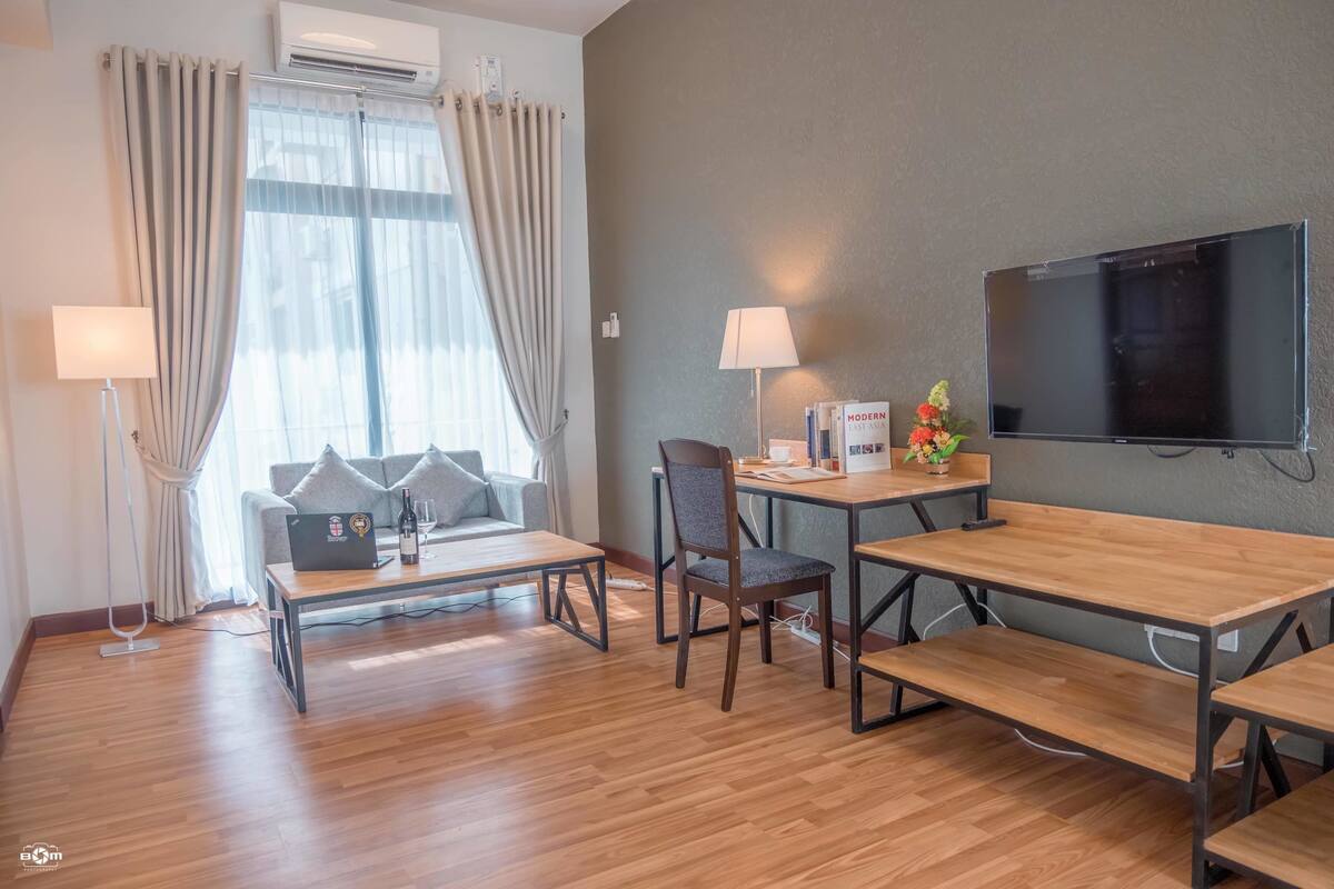Et hotelværelse med en sofa, et træbord, et skrivebord med stol, et fjernsyn og et vindue med gardiner.
