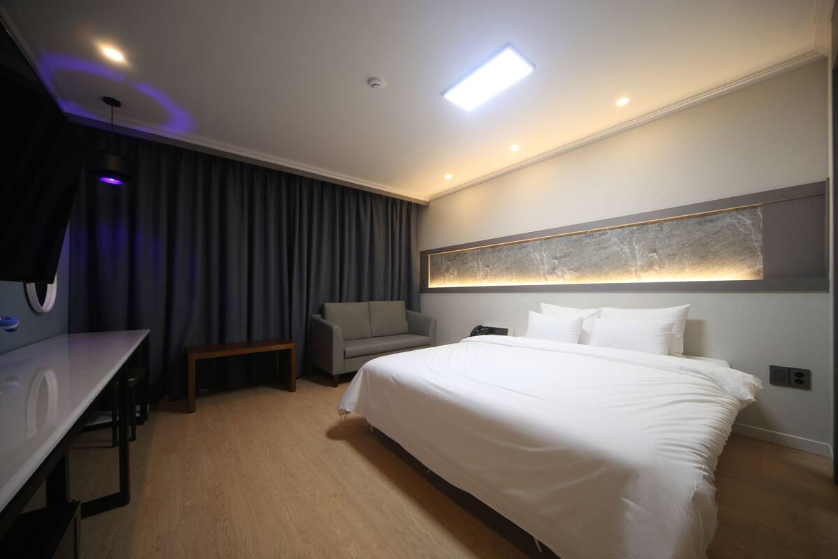 A modern hotel room with a large bed, a desk, a sofa, and a wall-mounted light fixture.