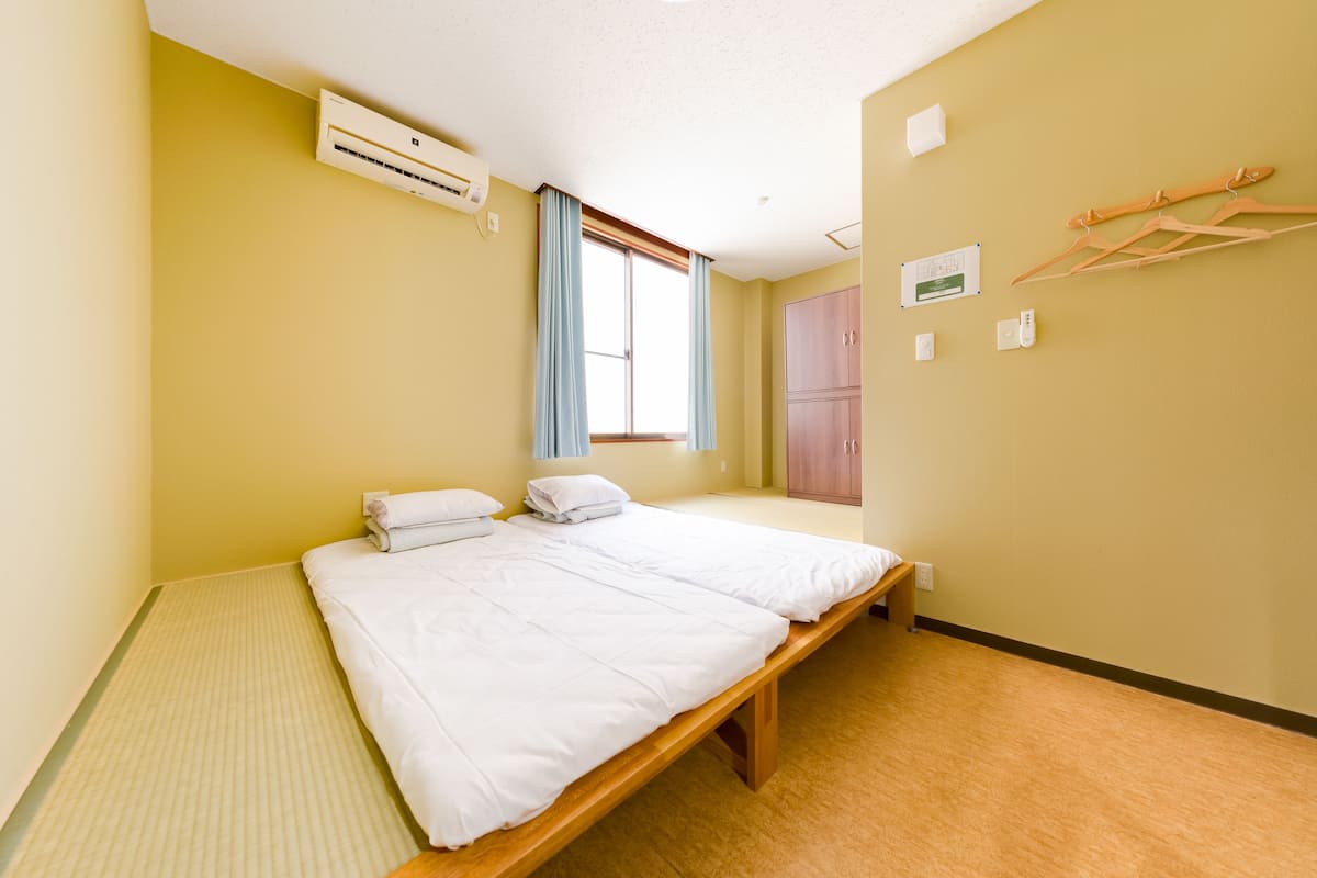 A single-bed room with a wooden bed frame, a window with curtains, an air conditioning unit, a wardrobe, and a wall-mounted drying rack.