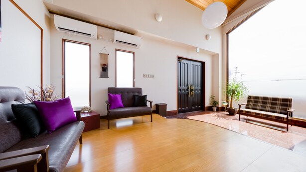 A spacious living room with a wooden floor, a sofa with purple cushions, a bench, and a potted plant. - Yolee Guest House (Izumisano)