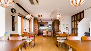 A dining area with wooden tables and chairs, a kitchen in the background, and pendant lighting. - Yolee Guest House (Izumisano)