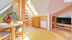 Ett modernt sovrum i loftstil med ett sluttande tak, takfönster, möbler i trä, en platt-TV och en matplats med bord och stolar.