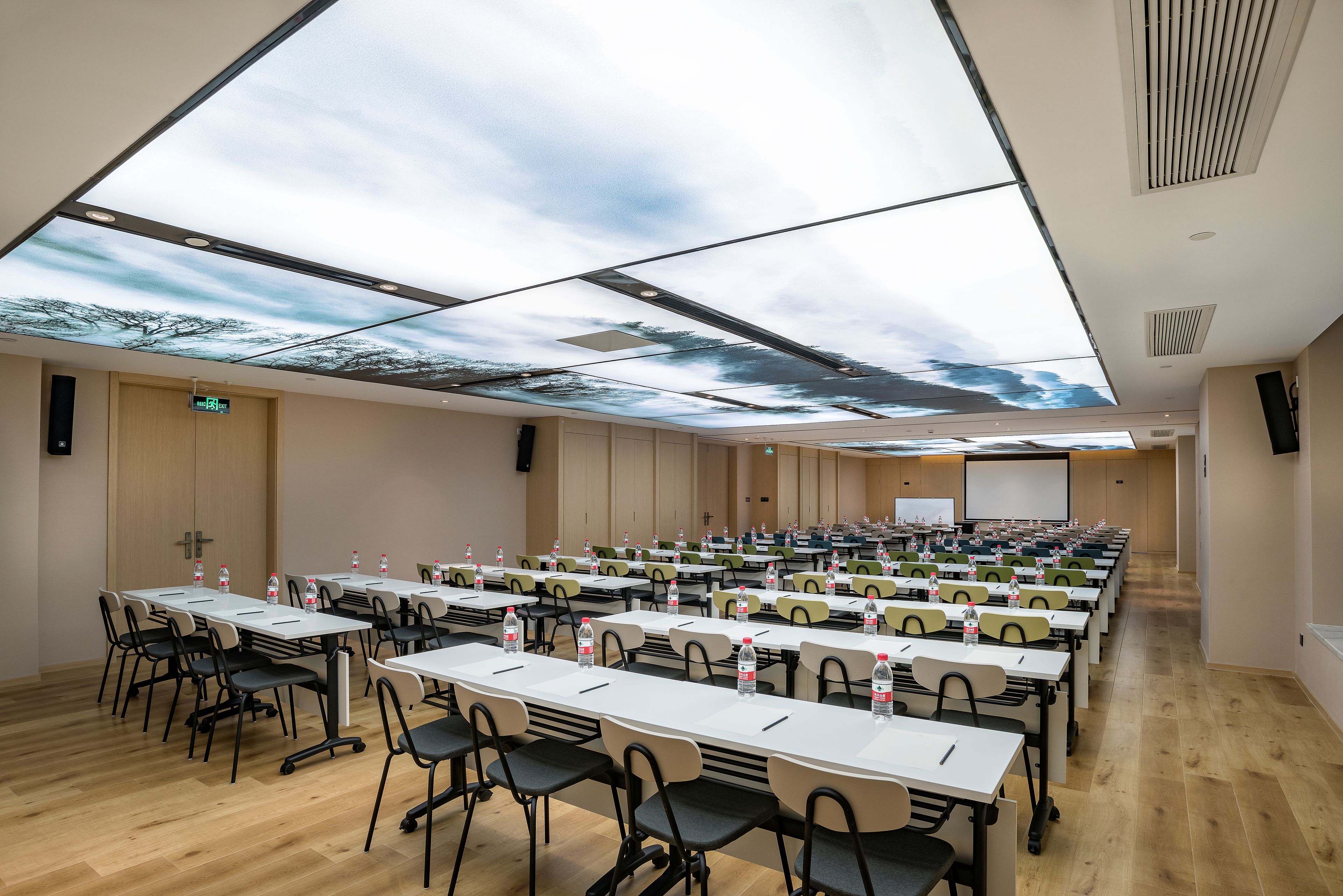 A conference room with rows of tables and chairs, a large projection screen, and a ceiling with a tree and sky design.