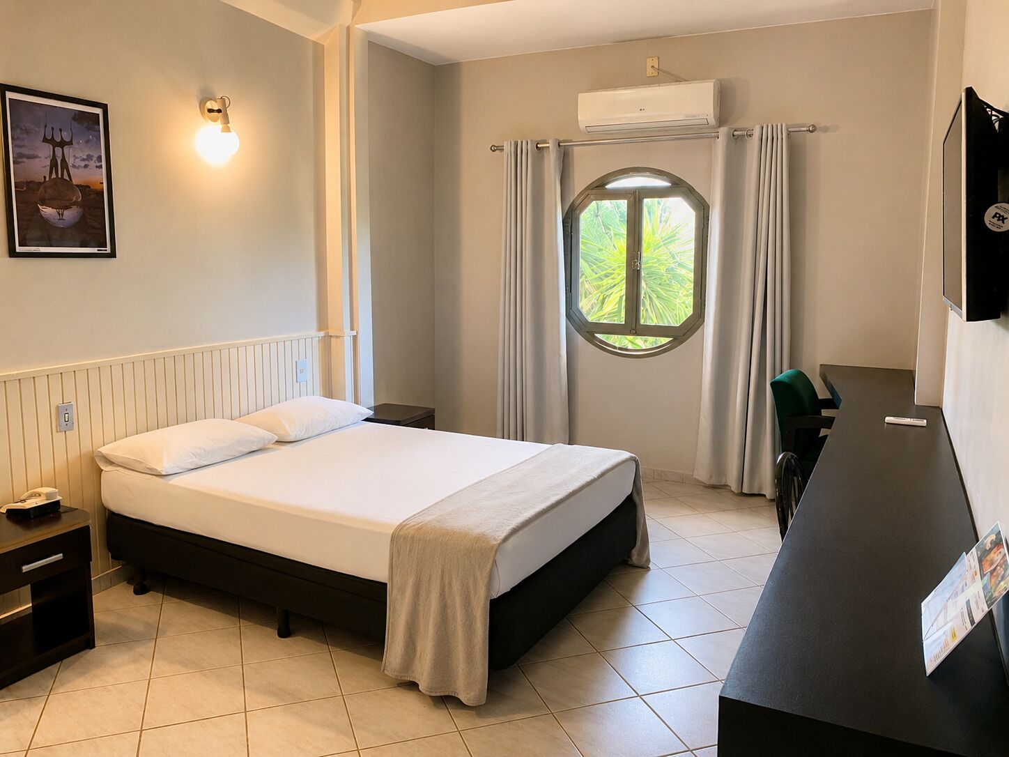 A hotel room with a bed, bedside table, lamp, window with curtains, air conditioning unit, and a desk with a chair.