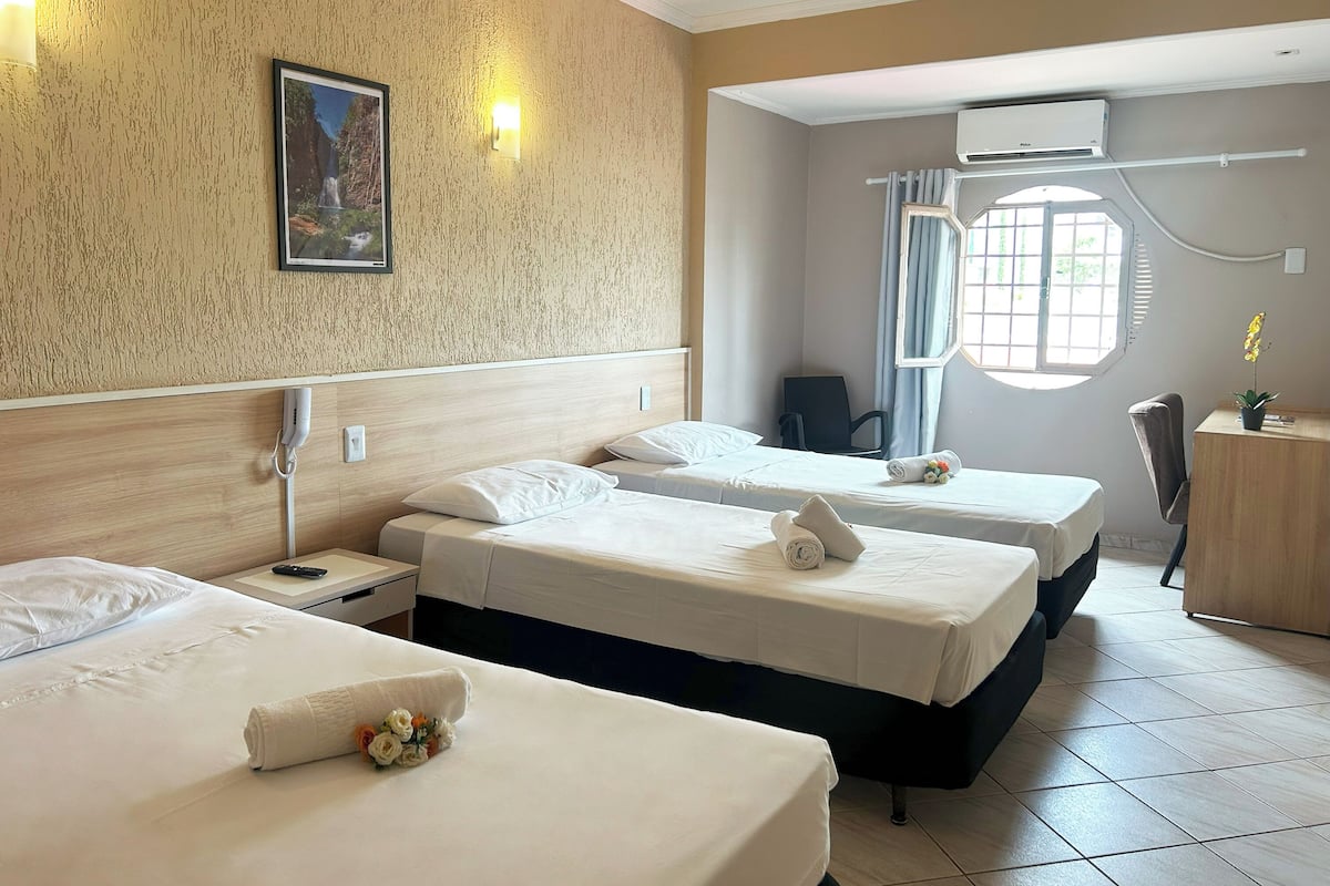 Ett hotellrum med tre sängar, ett skrivbord, en stol, en luftkonditioneringsanläggning och ett fönster med gardiner.