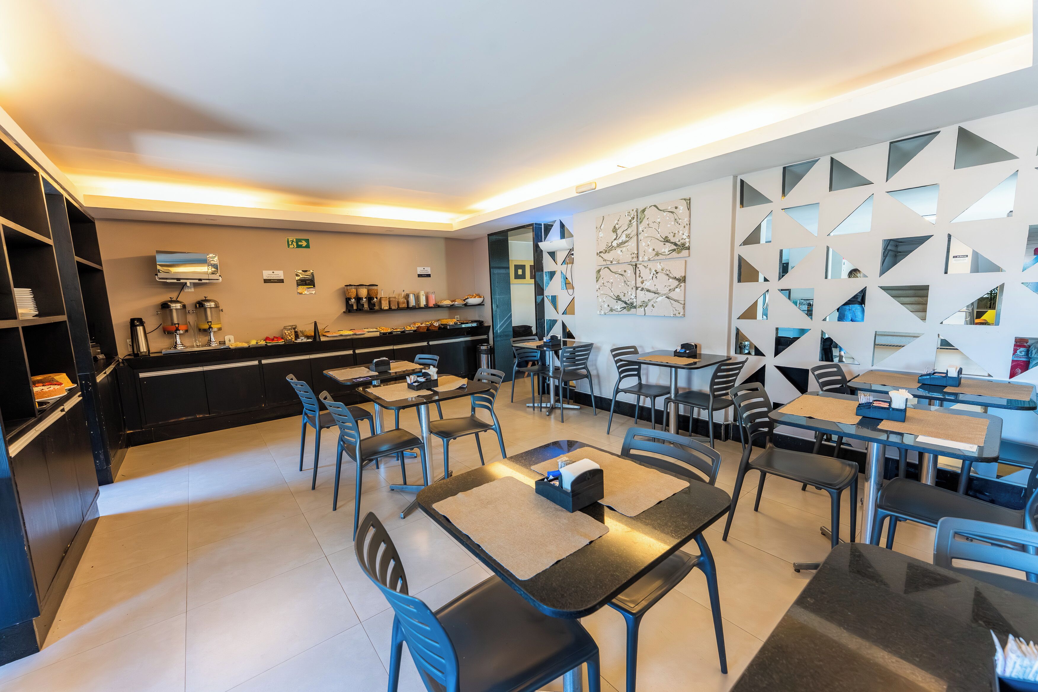 Un comedor moderno con mesas y sillas, una cocina al fondo y un cuadro de arte con motivos geométricos.