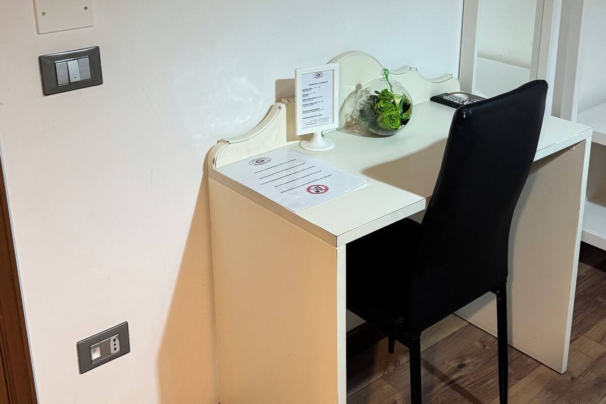 A small, white desk with a black chair, a glass vase with green plants, and a mirror on the wall.