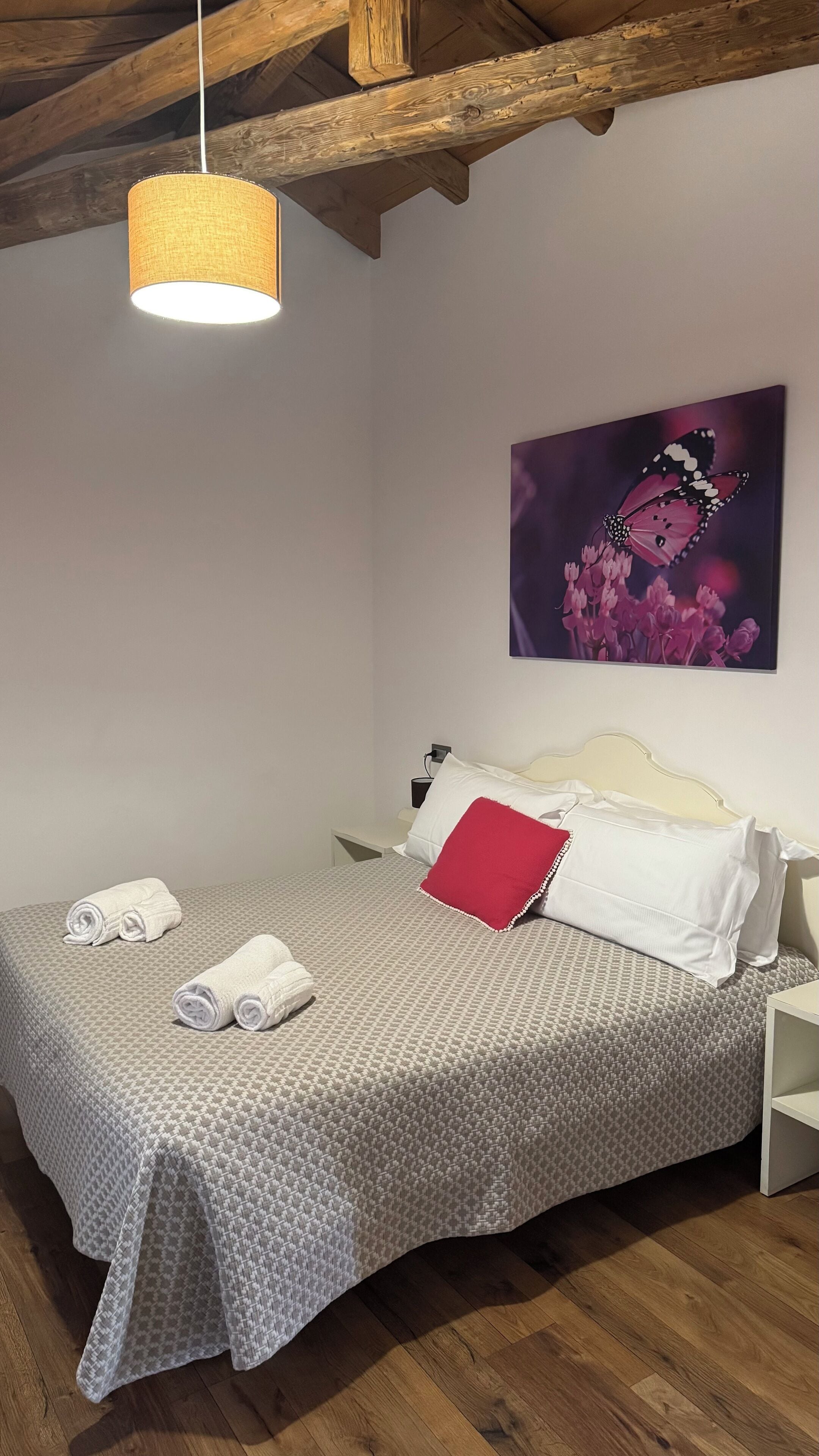 Cama bem arrumada com toalhas enroladas, um travesseiro vermelho e um quadro de uma borboleta na parede.