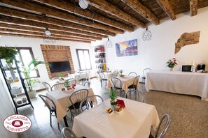 Un restaurante con vigas de madera a la vista, mesas y sillas blancas. Hay plantas en macetas y una pared de ladrillo.