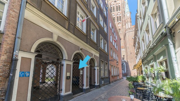 A narrow cobblestone street with buildings on either side, featuring arched doorways and a flag. - Apartament Przytulny –2 sypialnie w centrum Starówki Gdańskiej (Gdansk)
