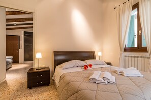 A neatly made bed with a beige quilt, two bedside lamps, and a nightstand with a lamp. - Corderie de la Tana Biennale Exhibition (Venice)