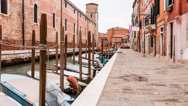 A narrow canal with boats docked on the left, a stone pathway on the right, and buildings with arched windows and balconies. - Corderie de la Tana Biennale Exhibition (Venice)
