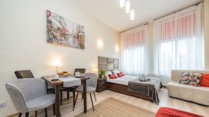 A modern hotel room with a dining area, a bed with a grey headboard, a sofa with a floral pillow, and a large window with sheer curtains. - Apartament Przytulny – centrum Starówki Gdańskiej (Gdansk)
