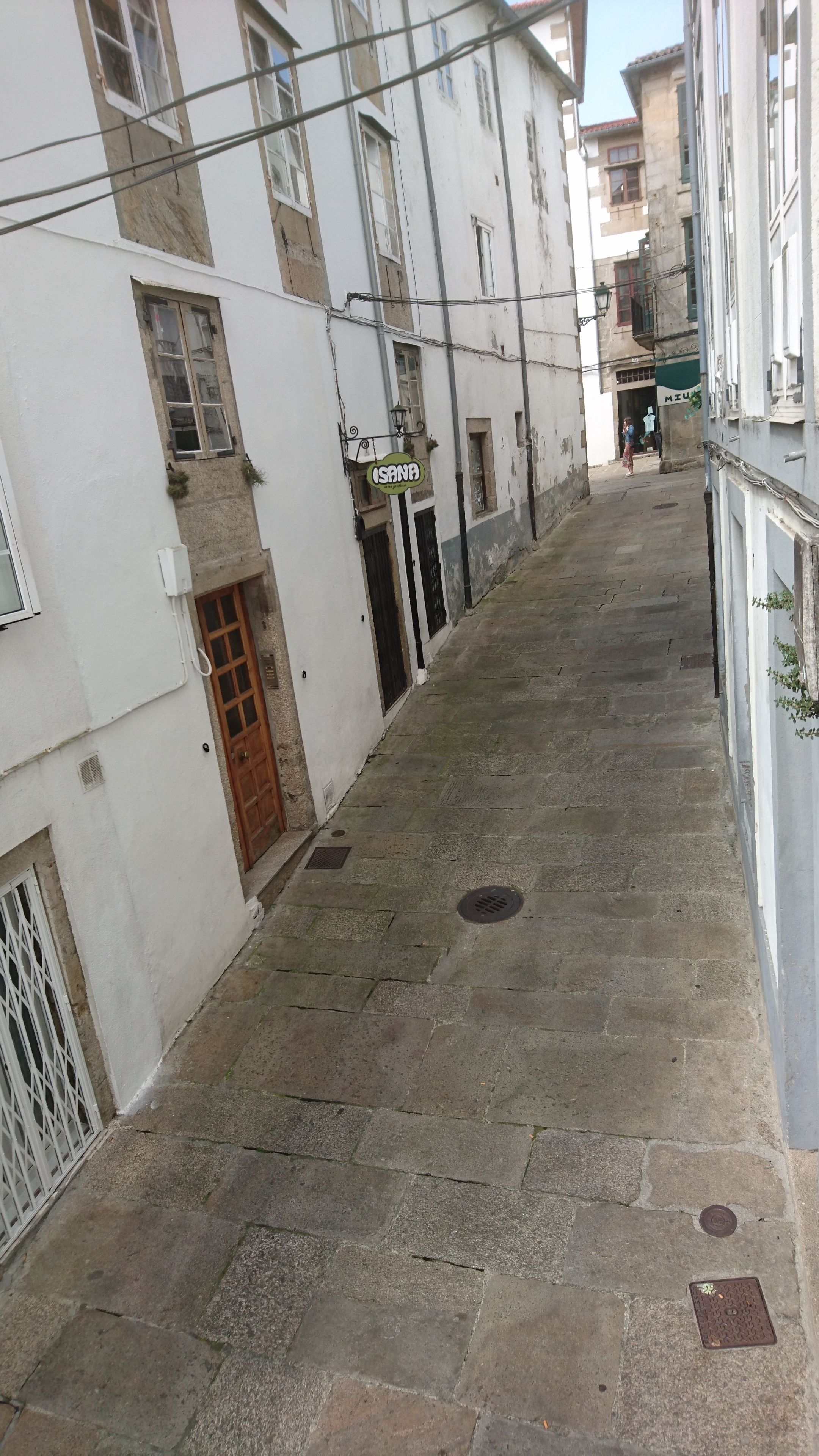 A narrow street with stone pavement, white buildings with multiple windows, and a visible door on the left.