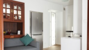 A compact living space with a grey sofa, a wooden cabinet, a white washing machine, and a door leading to another room. - Los truques (Santiago de Compostela)