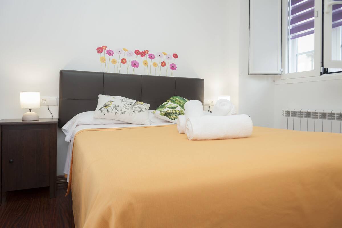 A neatly made bed with a yellow bedspread, a pillow with a floral pattern, and a rolled white towel on the side.