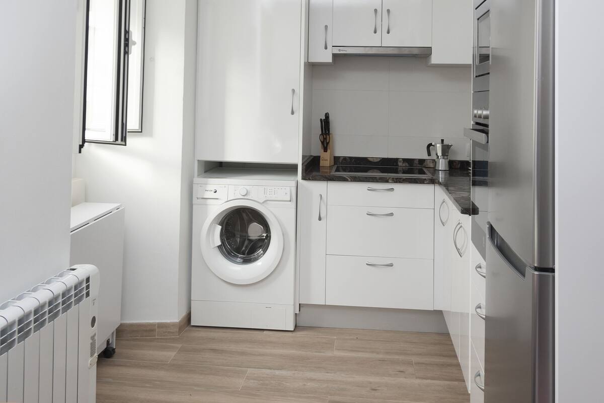 A compact kitchen with white cabinets, a built-in washing machine, and a stainless steel refrigerator.