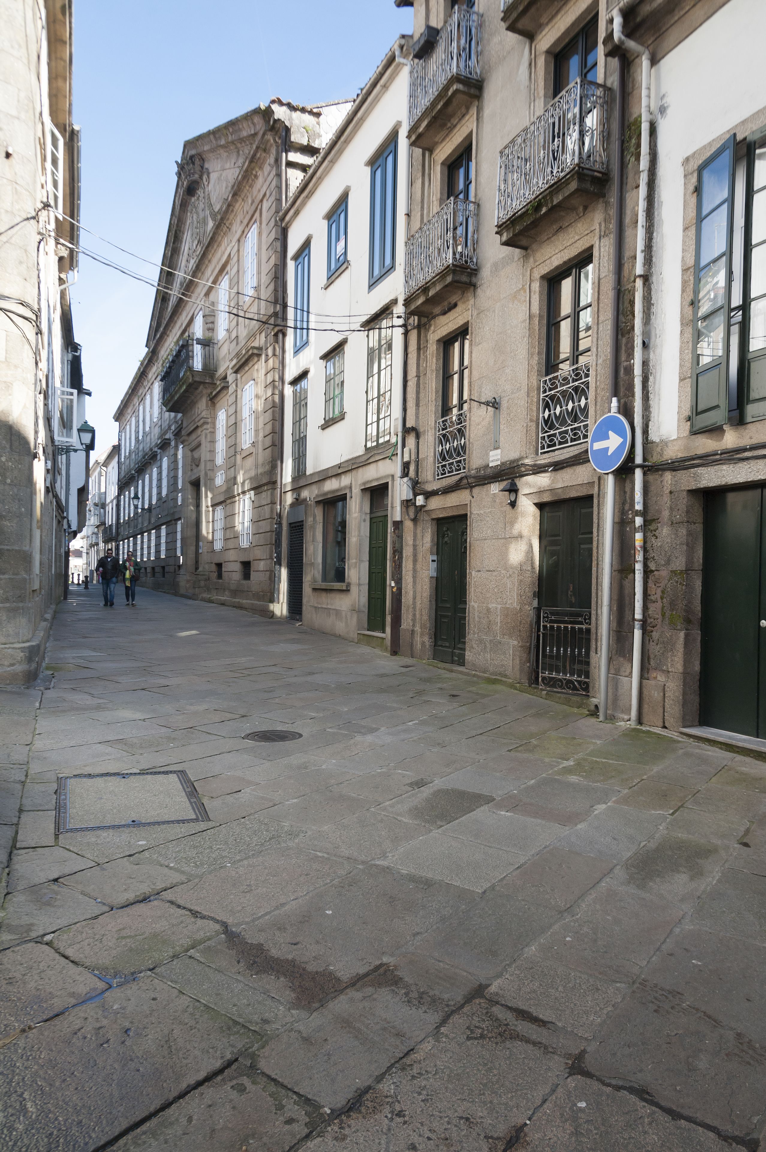 A narrow street lined with historic buildings, featuring stone facades, balconies, and a directional sign.