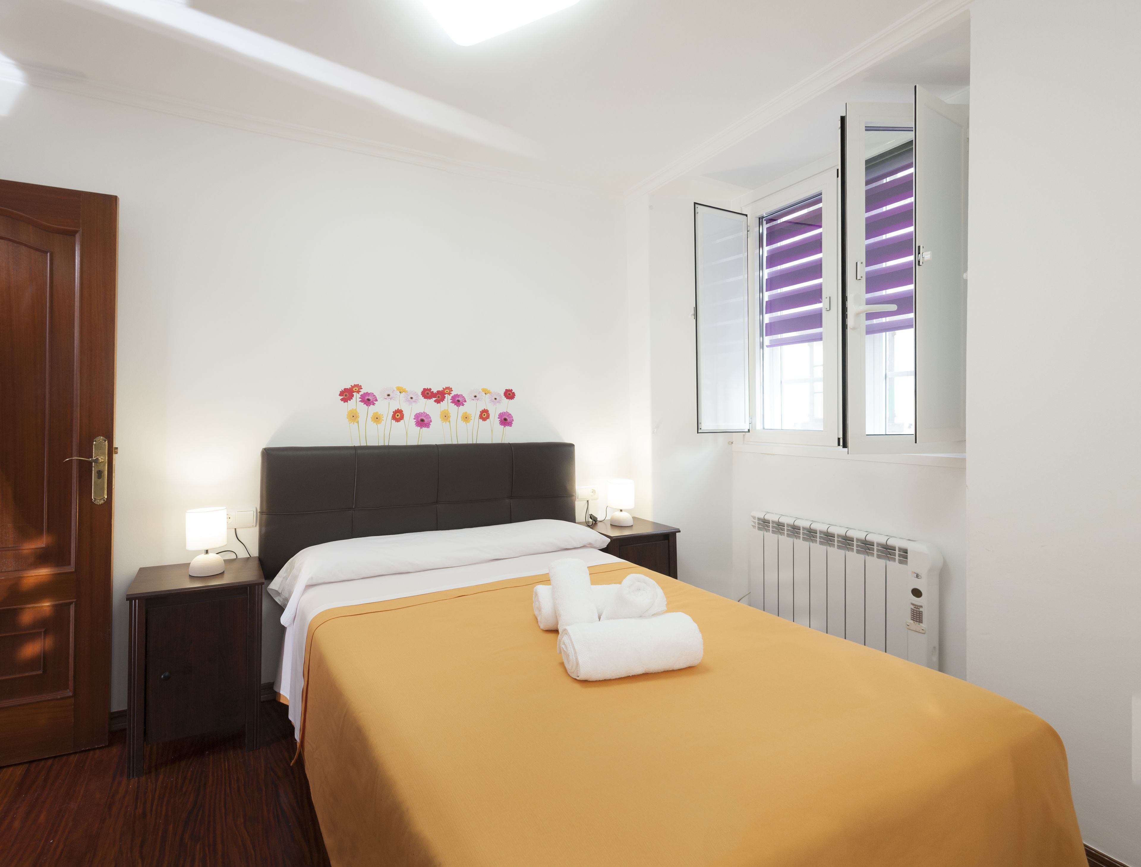 Habitación de hotel con cama, mesita de noche, adorno floral en la pared, ventana con persianas y radiador.